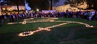 Een menigte verzamelde zich rond een groot, met kaarsen verlicht vredessymbool op een gazon in de schemering.