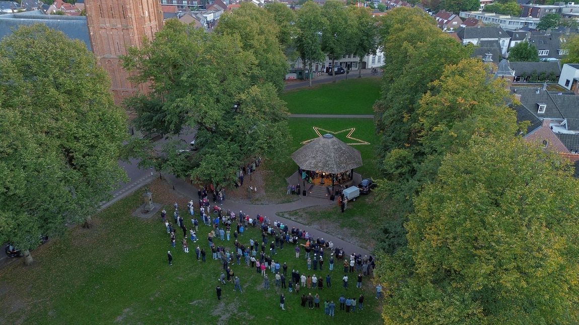Mensen hadden zich verzameld rond de kiosk op de Vrijthof in Hilvarenbeek, met bomen en stadsgebouwen in de buurt.