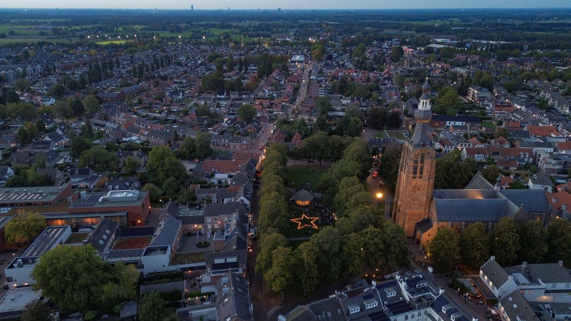 Luchtfoto bij schemering van een stad met dichte bebouwing, met bomen omzoomde straten en een verlichte kerktoren.