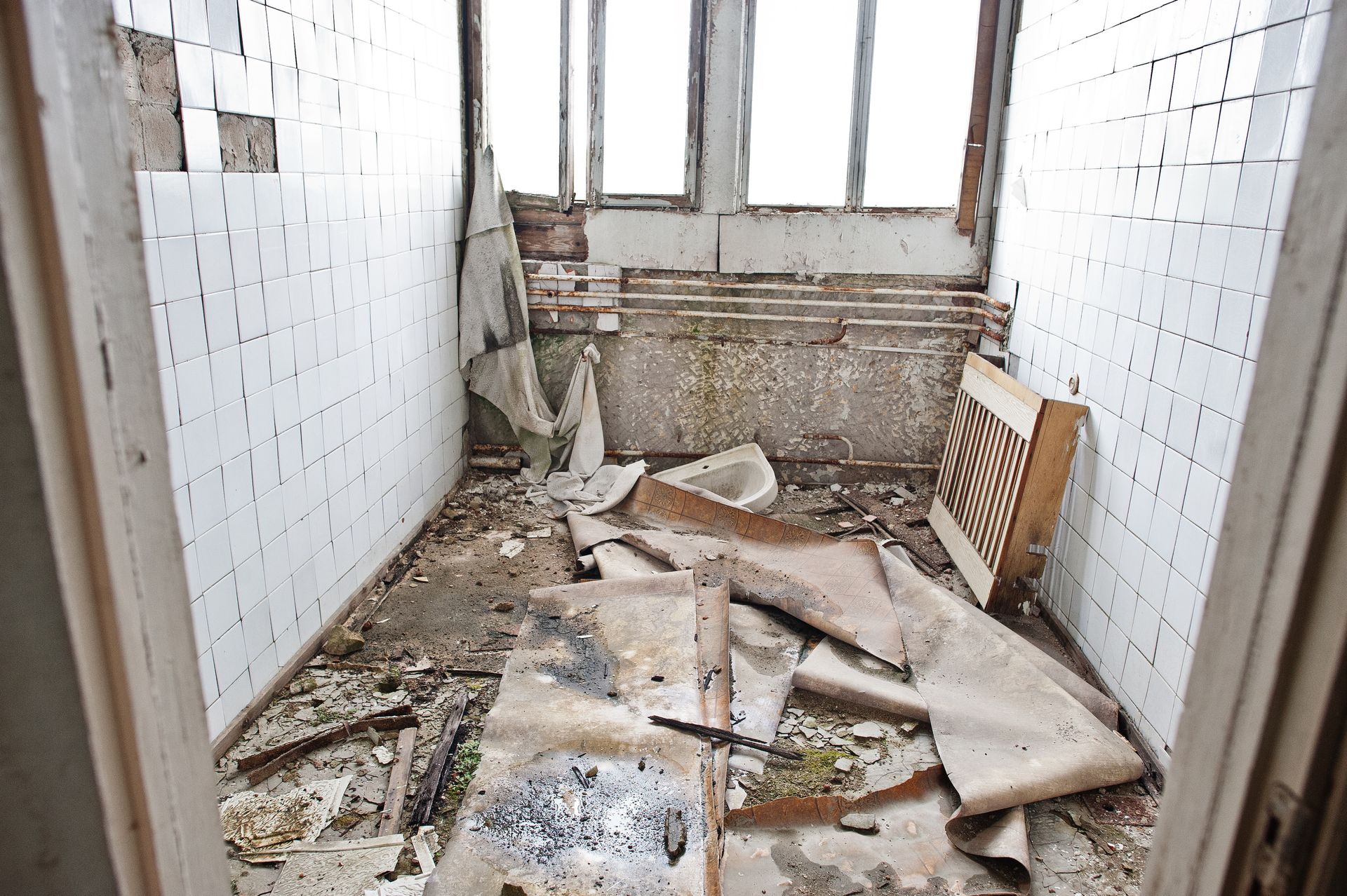 A dilapidated room with white-tiled walls, debris, and torn flooring under a window.