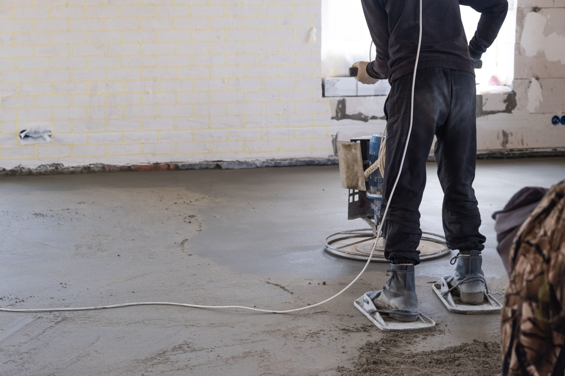 A worker in dark clothing uses a power trowel to smooth a wet concrete floor in an unfinished indoor room.