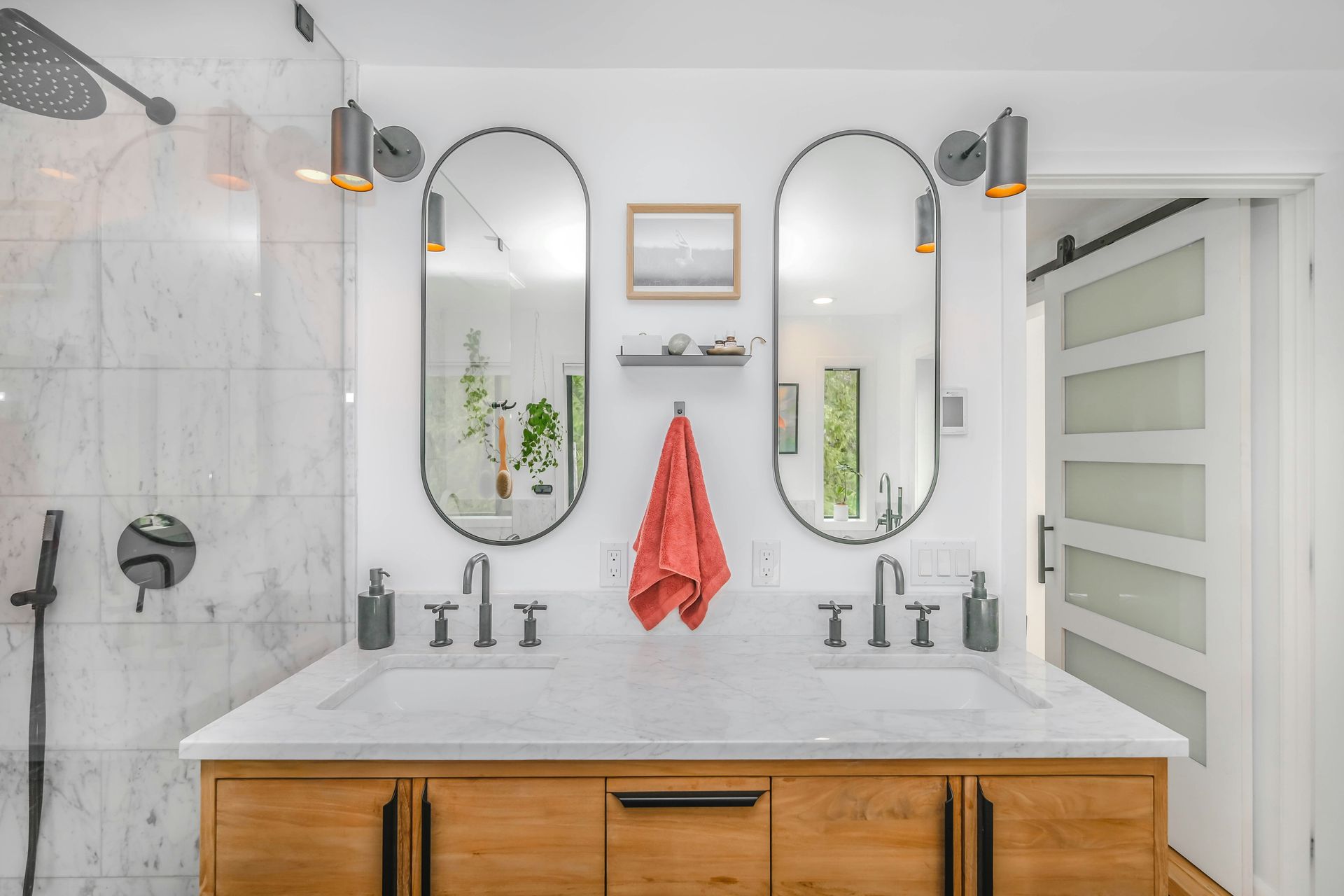 Modern bathroom vanity with a light wood cabinet, white marble countertop, dual sinks, oval mirrors, and a red towel.