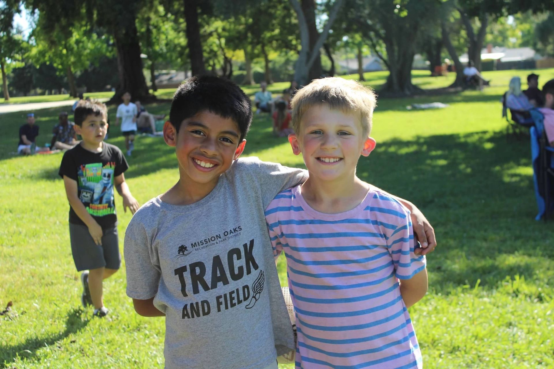 Two smiling boys with arms around each other in a park. One wears a track shirt, the other a striped shirt.