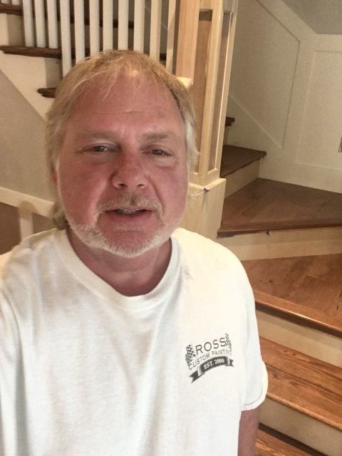 Man in a white T-shirt standing indoors by a wooden staircase, looking at the camera.