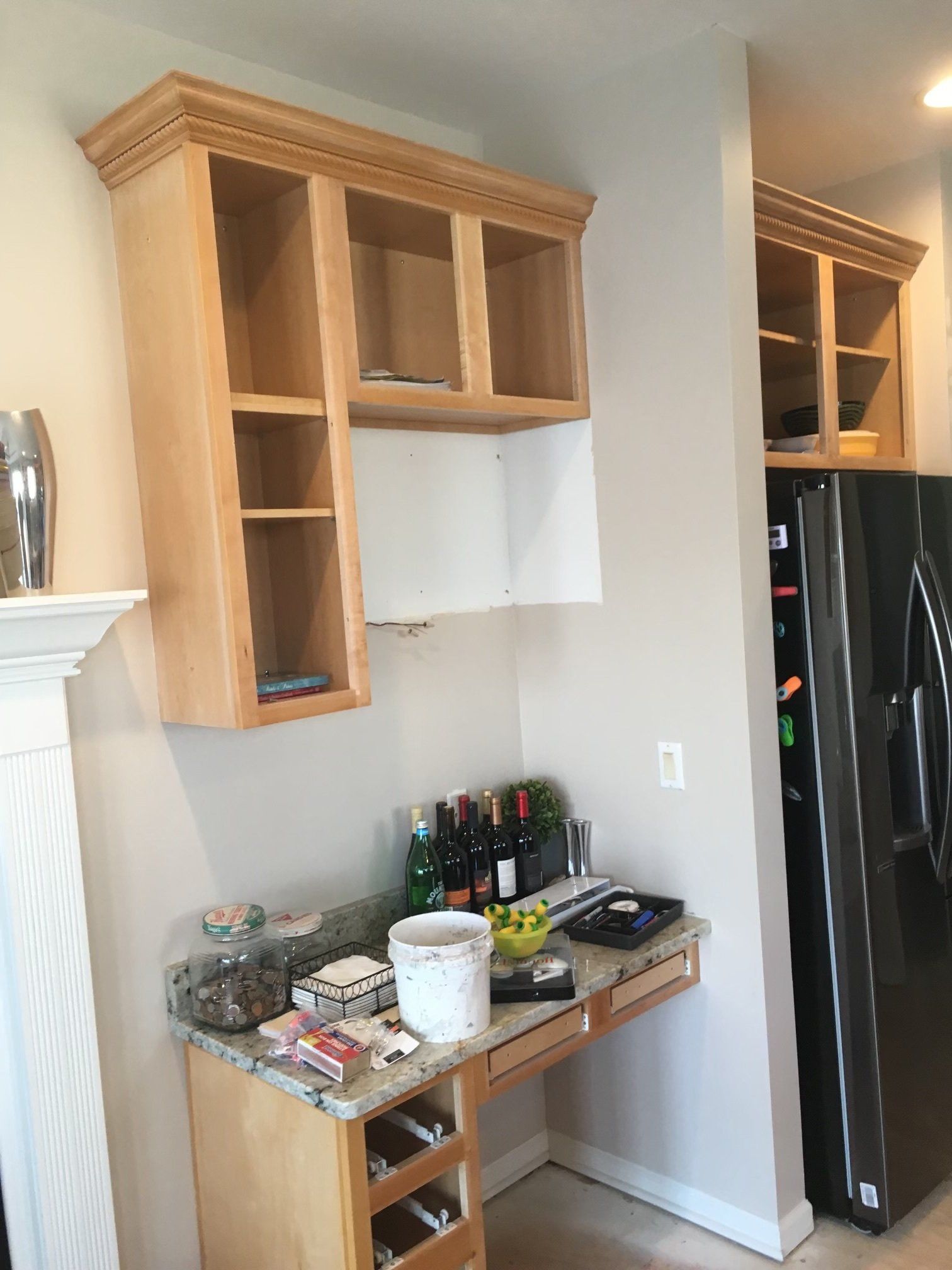Kitchen cabinet above a small counter with drinks, bowls, and utensils beside a black refrigerator.