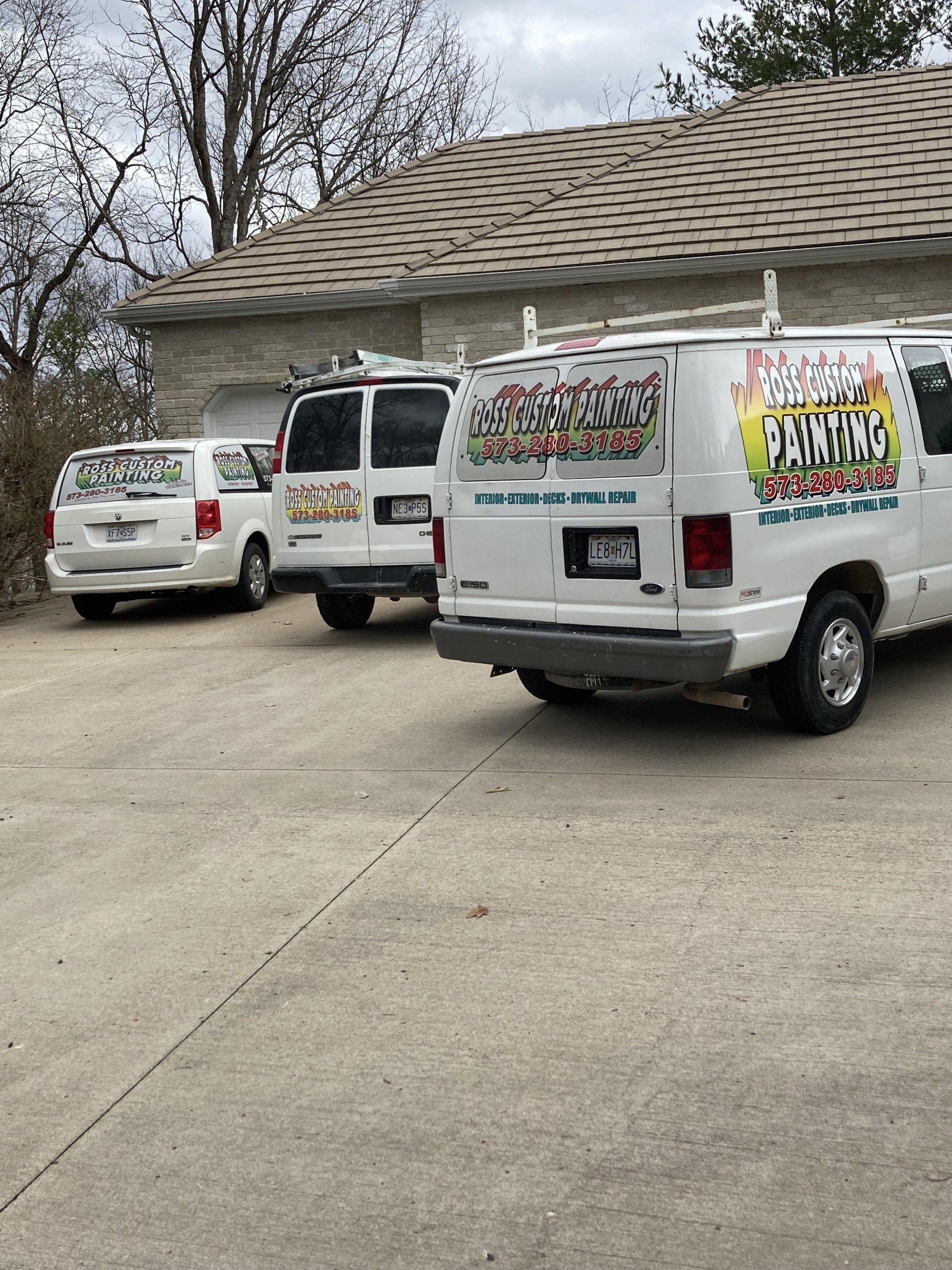 Three white work vans parked in a driveway, with a house and leafless trees in the background.