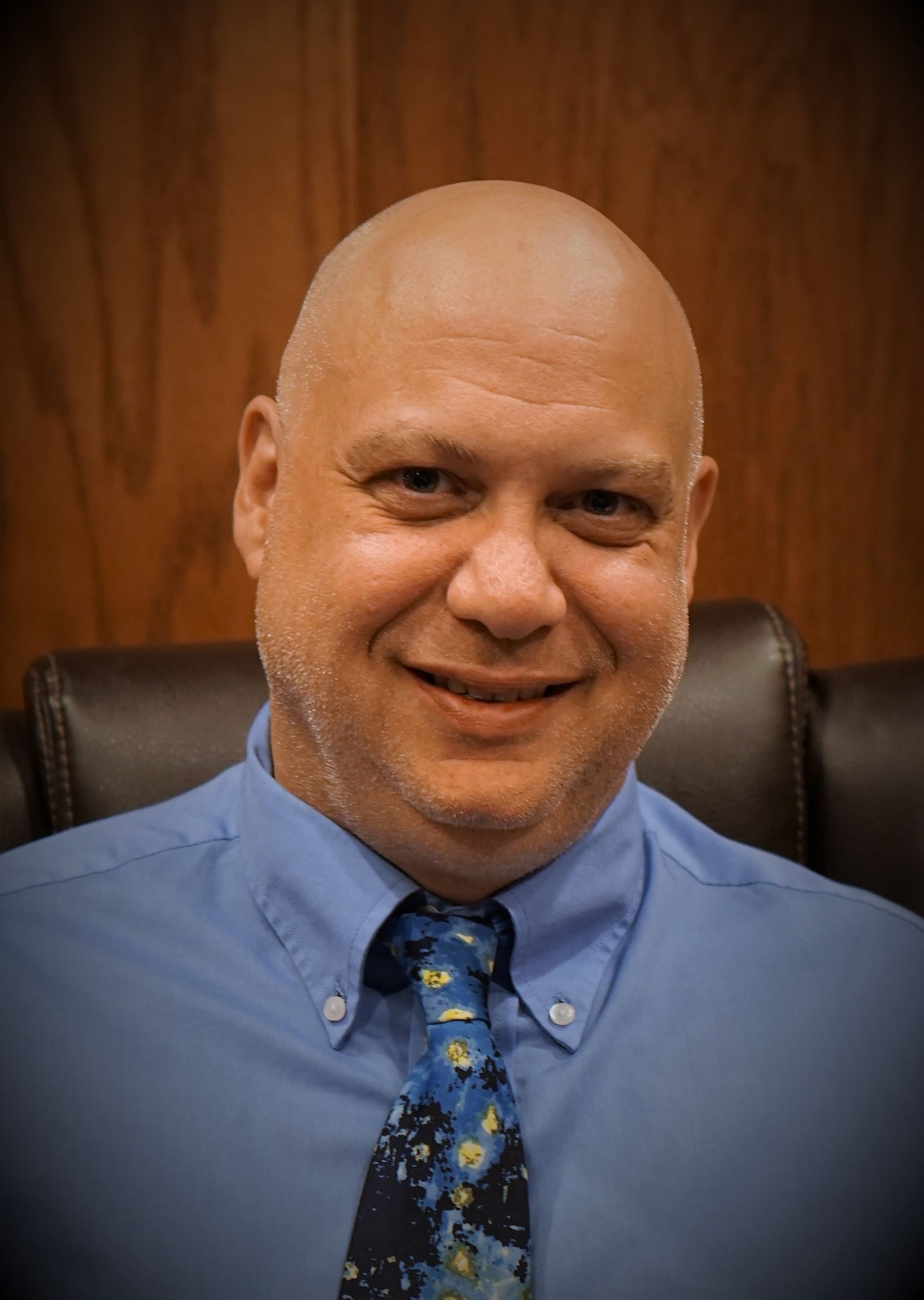 A bald man wearing a blue shirt and tie is smiling for the camera.