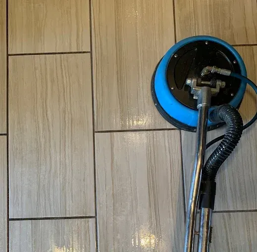 A professional floor-cleaning tool with a blue circular head operates on light-colored, wood-look rectangular floor tiles.