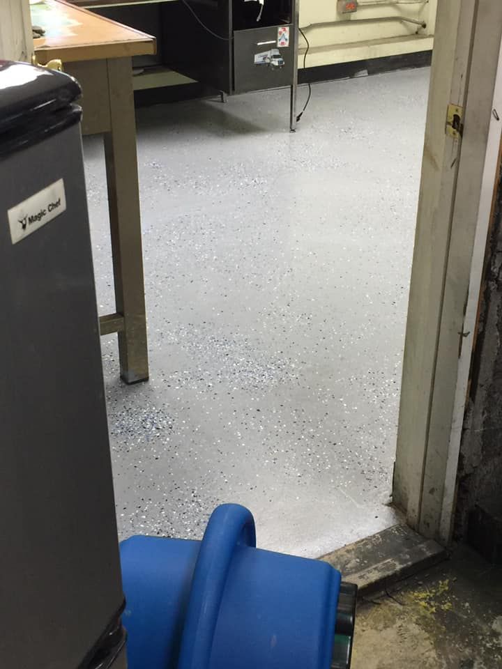 A gray speckled epoxy floor viewed through a doorway, with a blue industrial fan and part of a refrigerator in the frame.