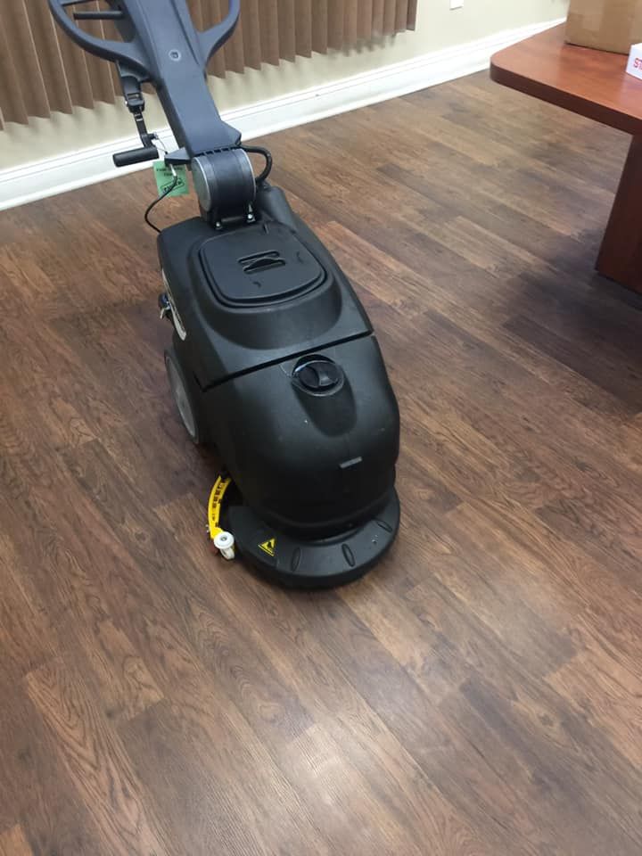 A black floor scrubber machine on a brown wood-look floor in an indoor office setting.