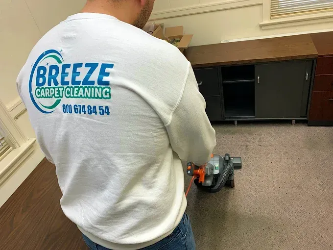 A person wearing a Breeze Carpet Cleaning shirt uses a machine to clean an office carpet near a dark cabinet.
