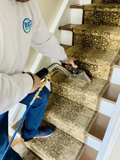 A technician in a white work shirt uses a handheld steam extraction wand to clean a patterned carpet on a staircase.