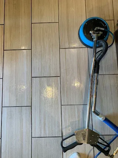 A tile floor cleaning machine with a blue circular head rests on wood-look ceramic tile flooring.