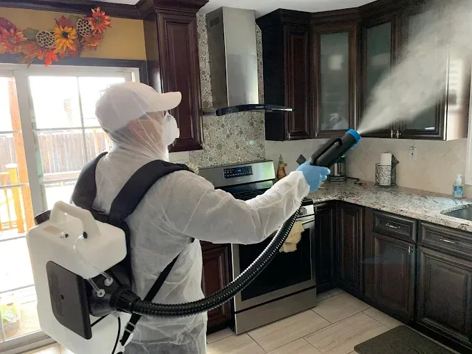 A professional in a protective white suit and mask uses a backpack sprayer to disinfect a kitchen.