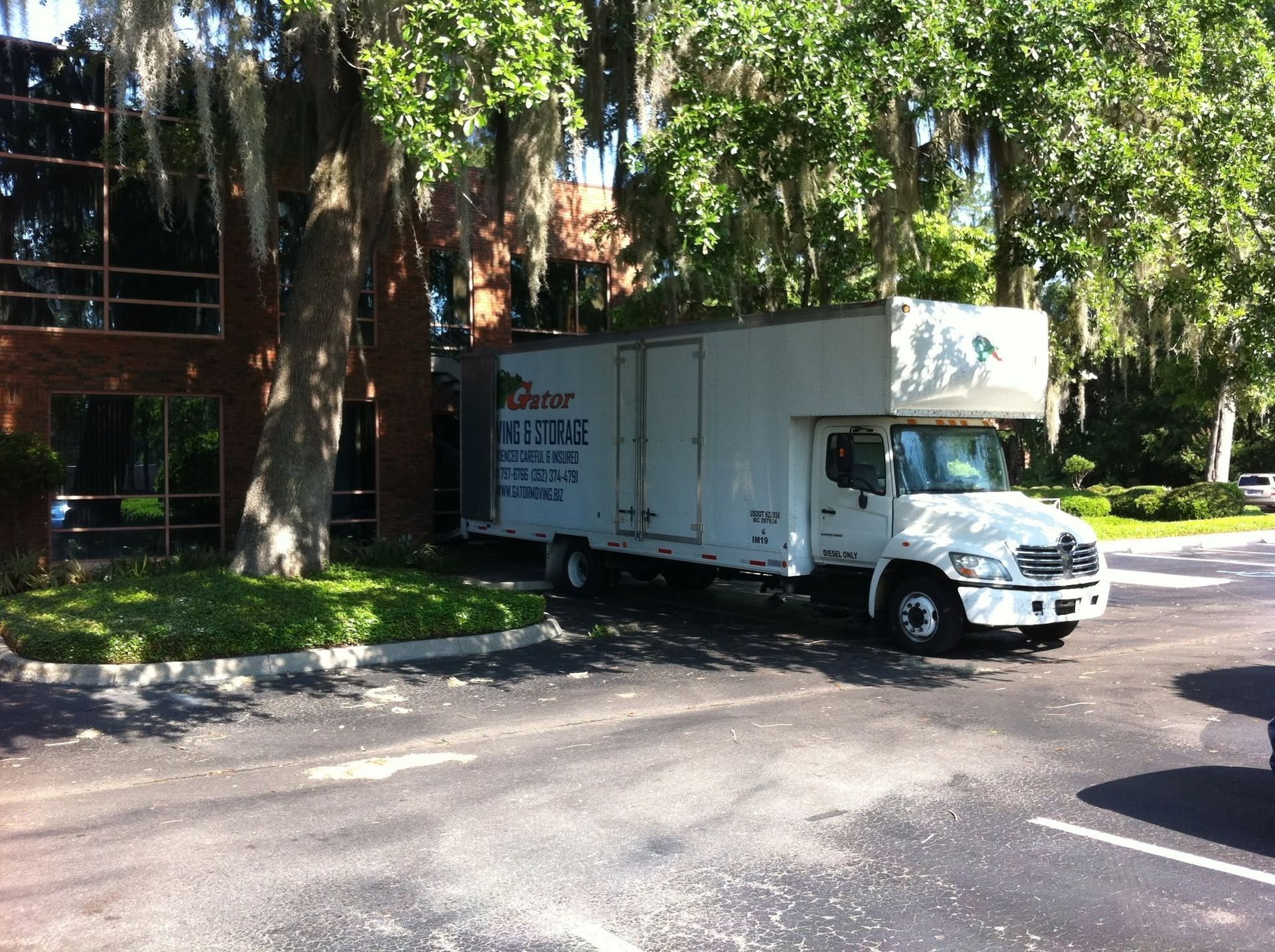 White delivery truck parked on a tree-lined street beside a building in sunlight
