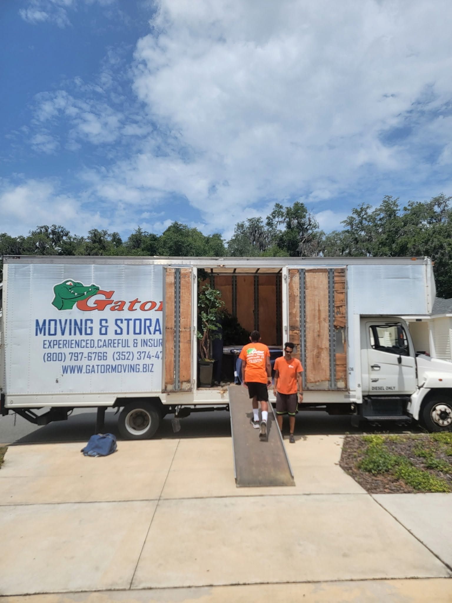 Moving truck with open side door and ramp; two workers in orange shirts loading items on a driveway.