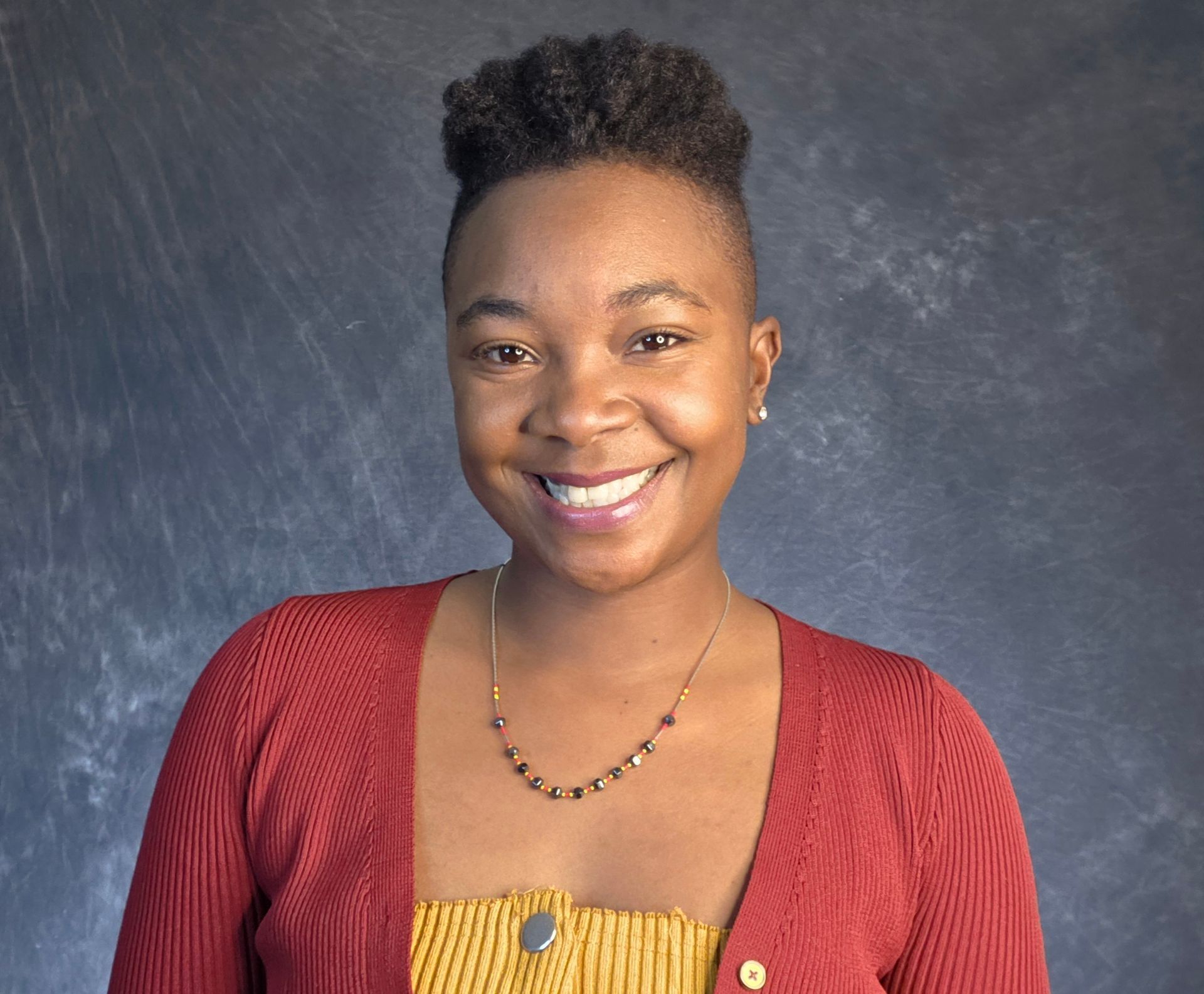 Smiling person in a red cardigan and yellow top against a blue-gray studio backdrop