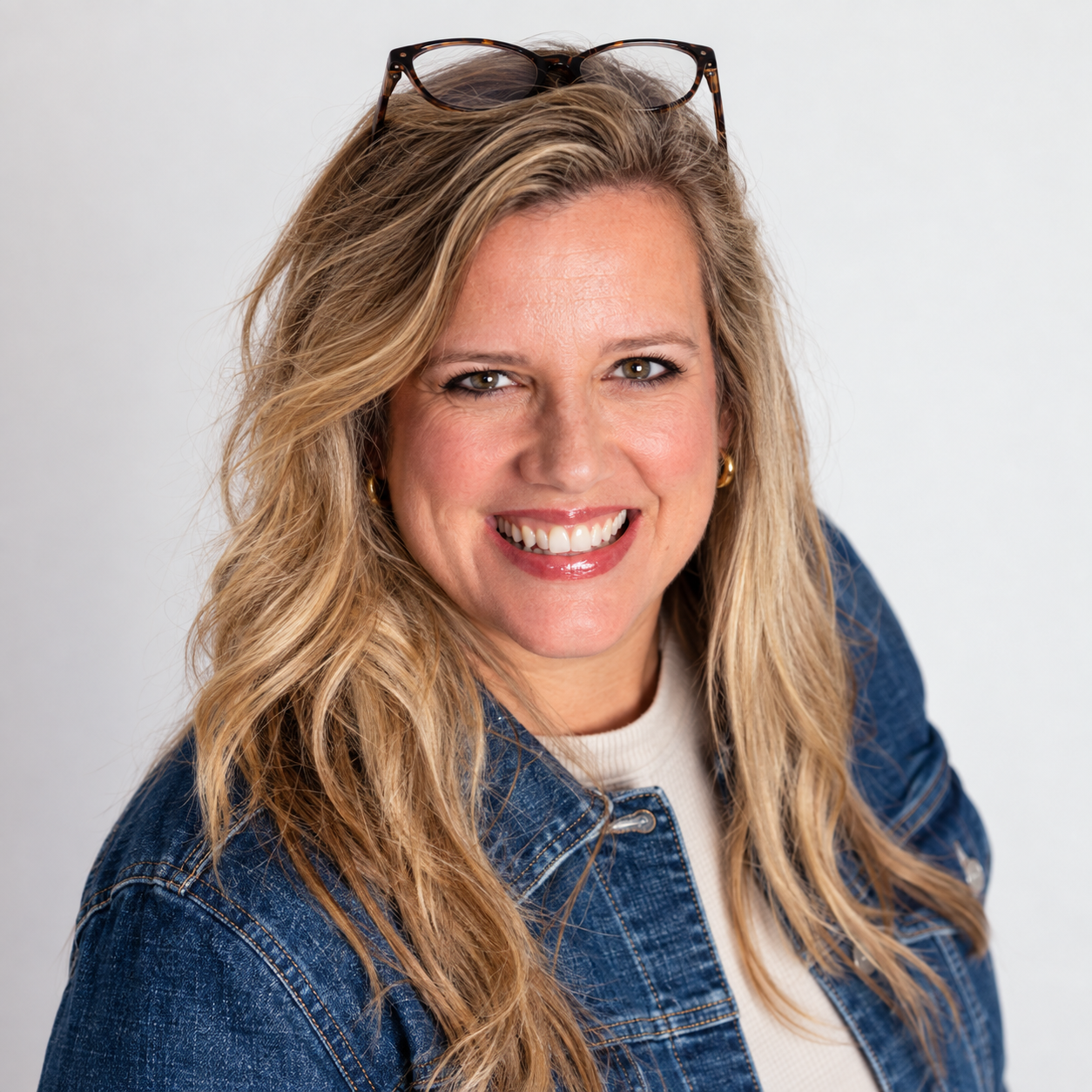Smiling woman with blonde hair in a denim jacket, wearing glasses on her head, against a light background