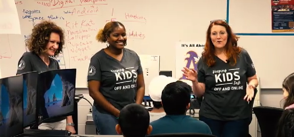 Three presenters in black “KIDS” shirts speaking in a classroom to an audience, with a whiteboard behind them