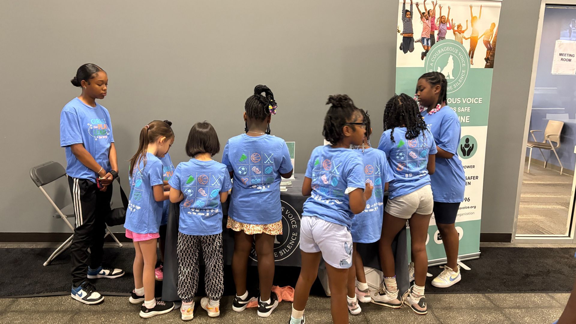 Group of children in blue shirts gathered around a display booth in a hallway.