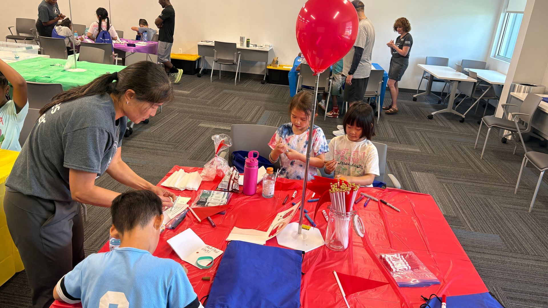 Children making crafts at a red-covered table with a large red balloon in a bright room.