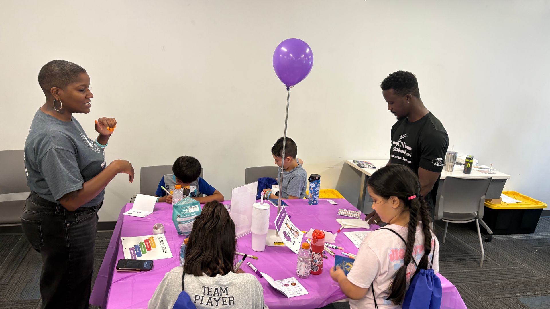 People gathered around a purple-decorated party table with a balloon in a room, chatting and eating snacks