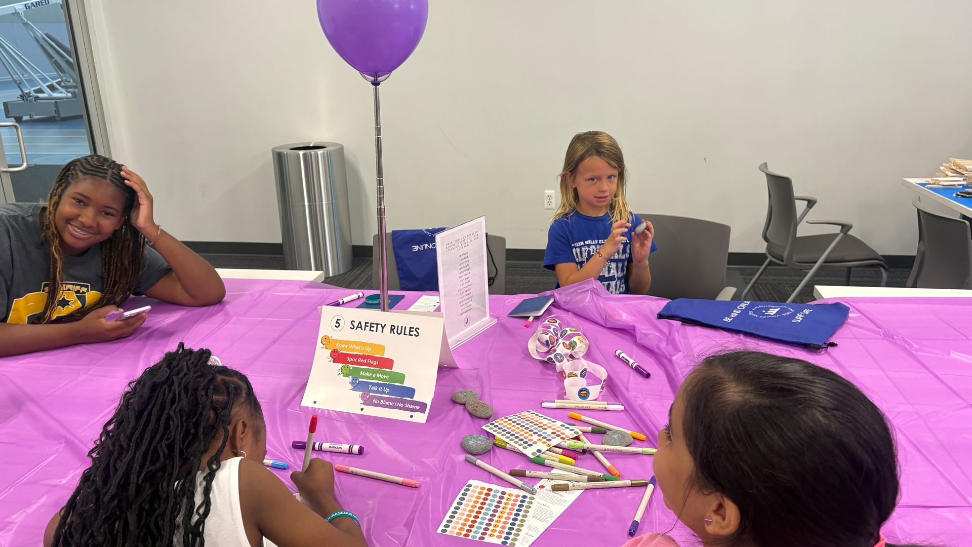 Children at a purple party table with balloons, crafts, and a “Happy Birthday” sign