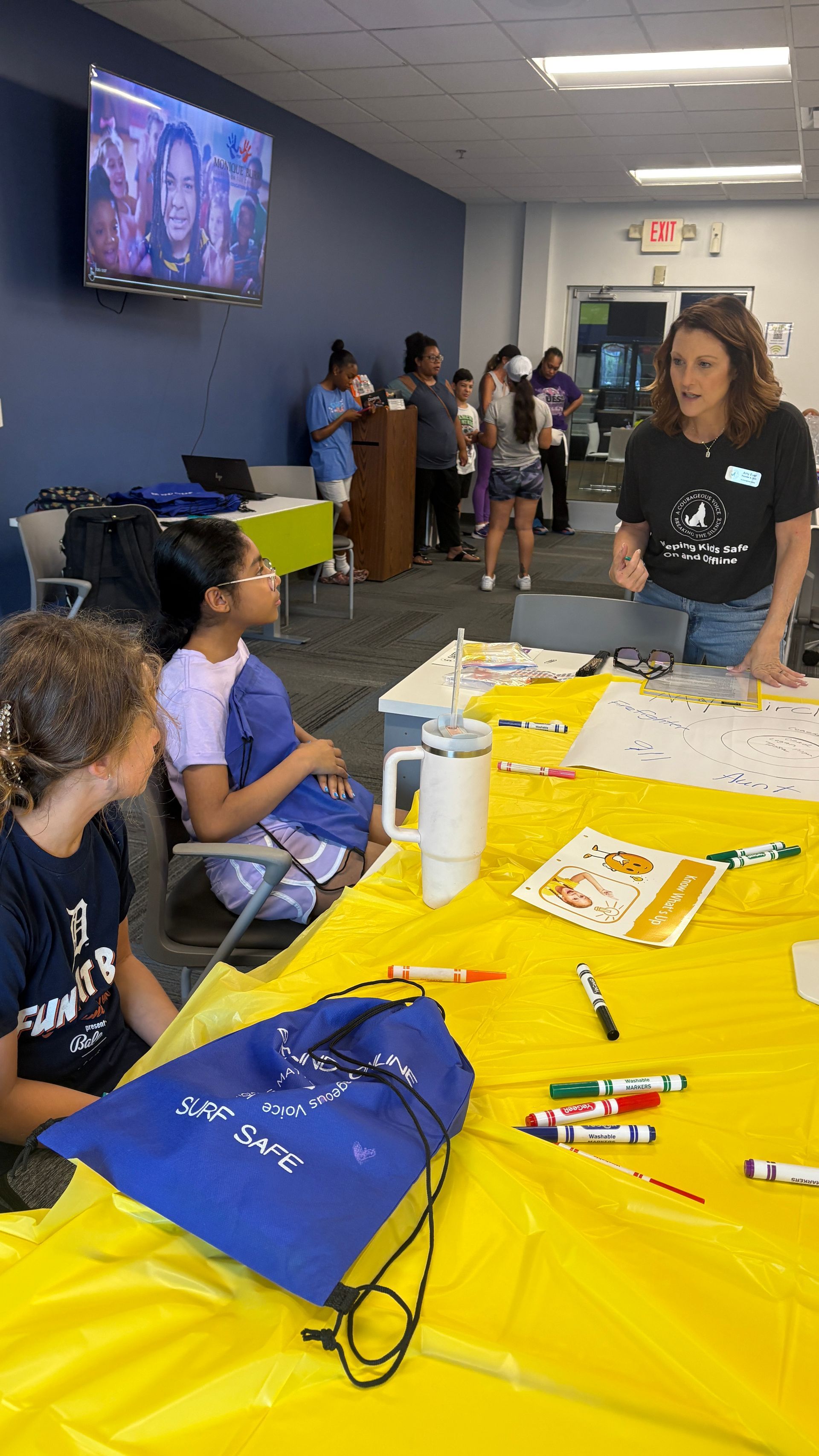 People at a yellow-covered table in a hallway, with crafts and supplies spread out during an indoor event.