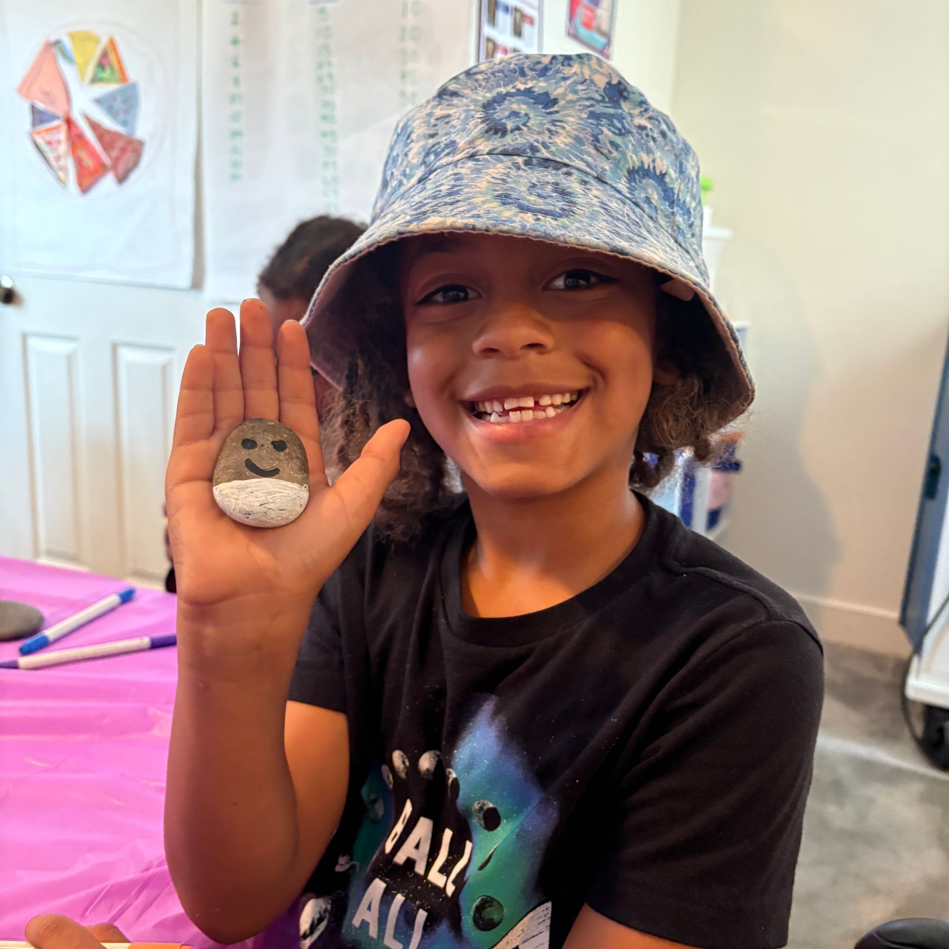 Smiling child in a blue patterned hat holds up a painted rock craft indoors.