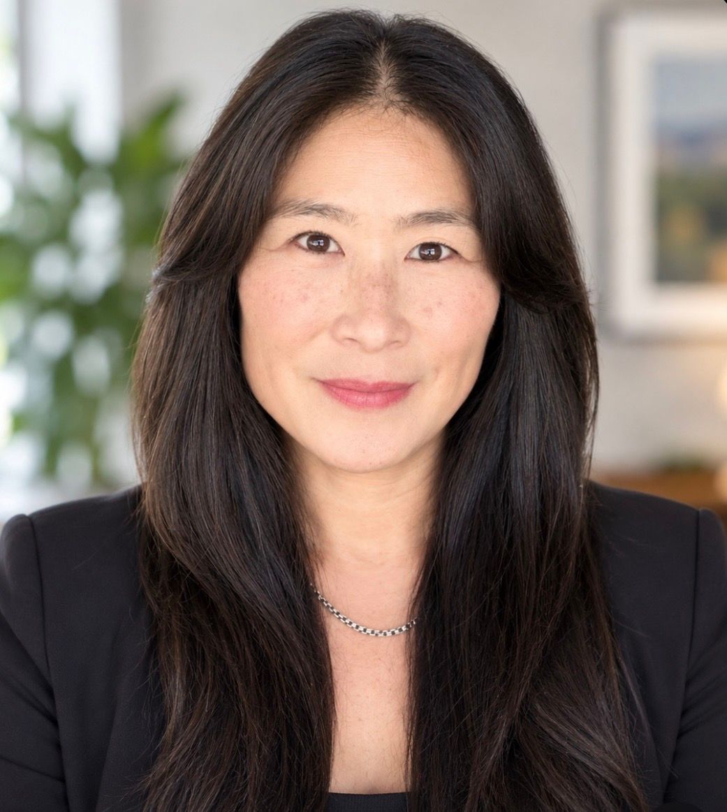Portrait of a woman with long dark hair, wearing a black blazer, against a softly blurred indoor background.