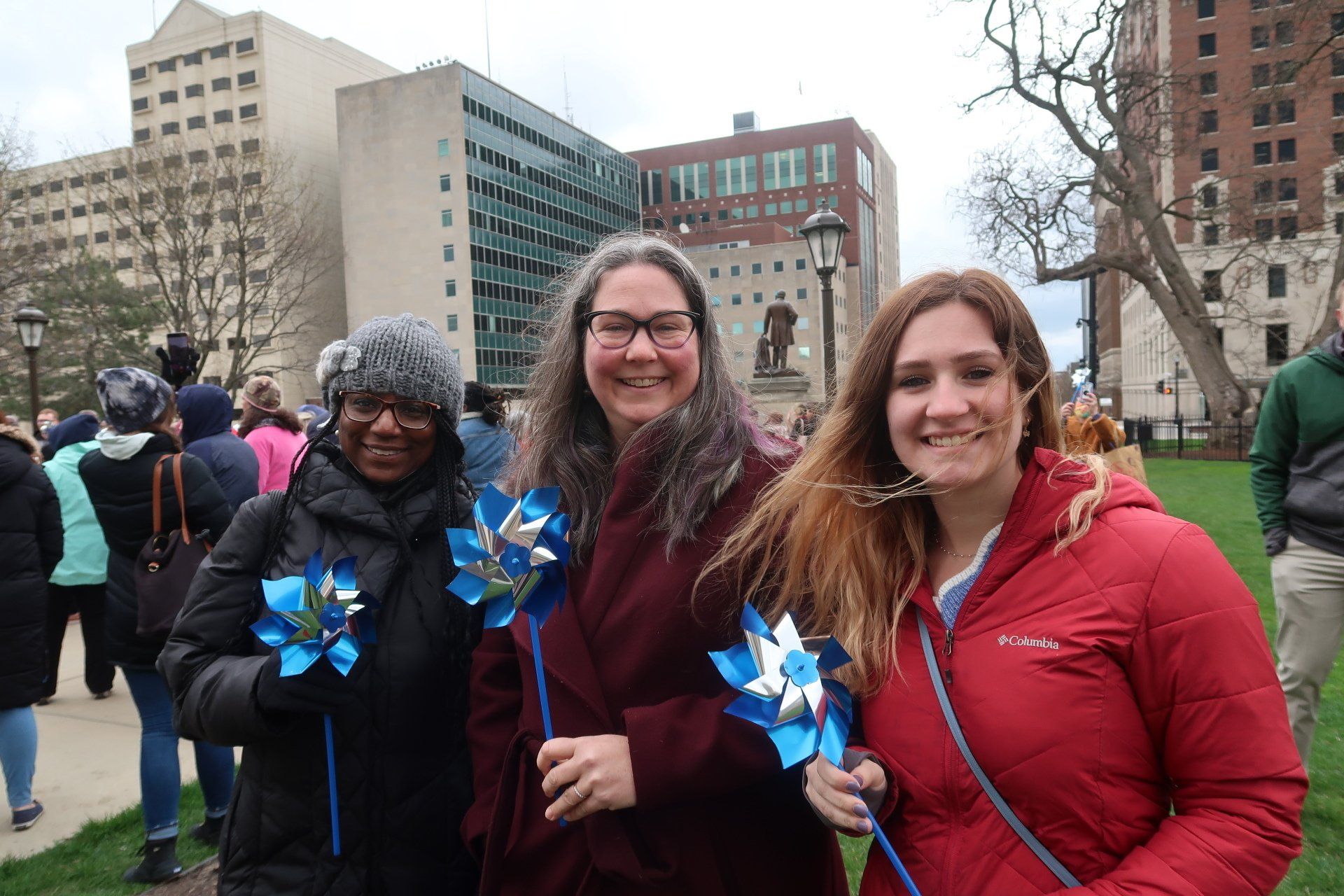Three people hold blue pinwheels, smiling outdoors near buildings.
