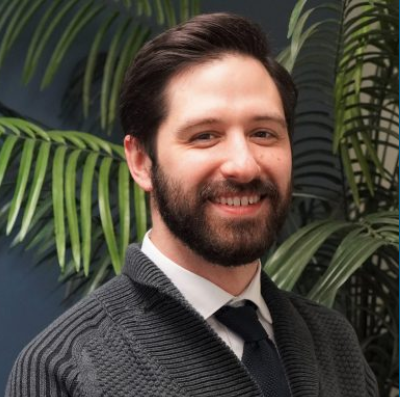 Smiling bearded person in a dark sweater and tie, seated indoors with palm plants behind them