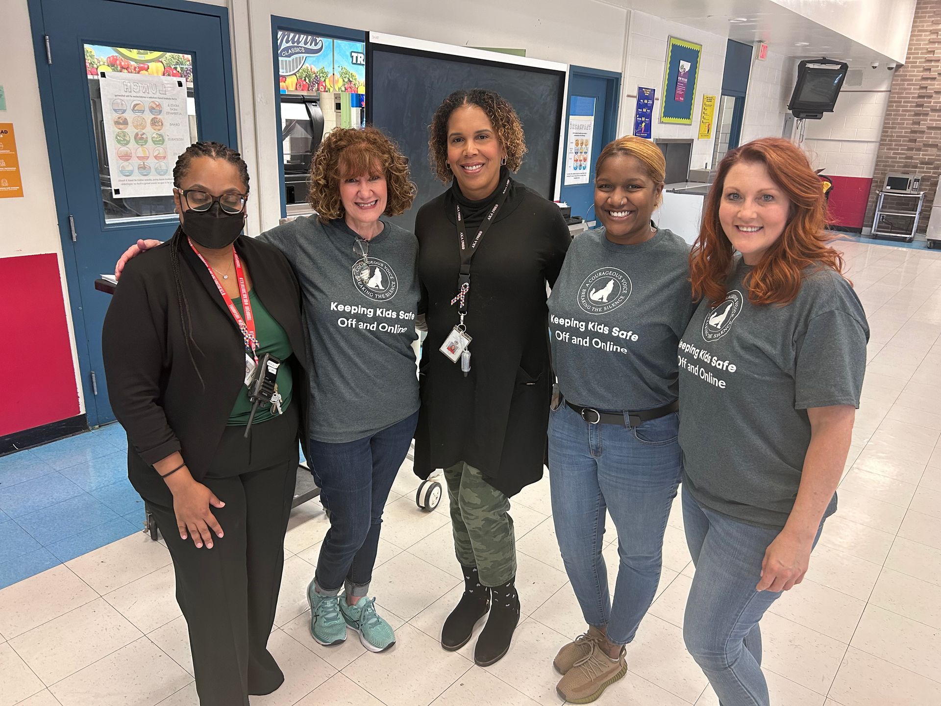 Five adults pose smiling in a school hallway, wearing dark shirts and jeans.