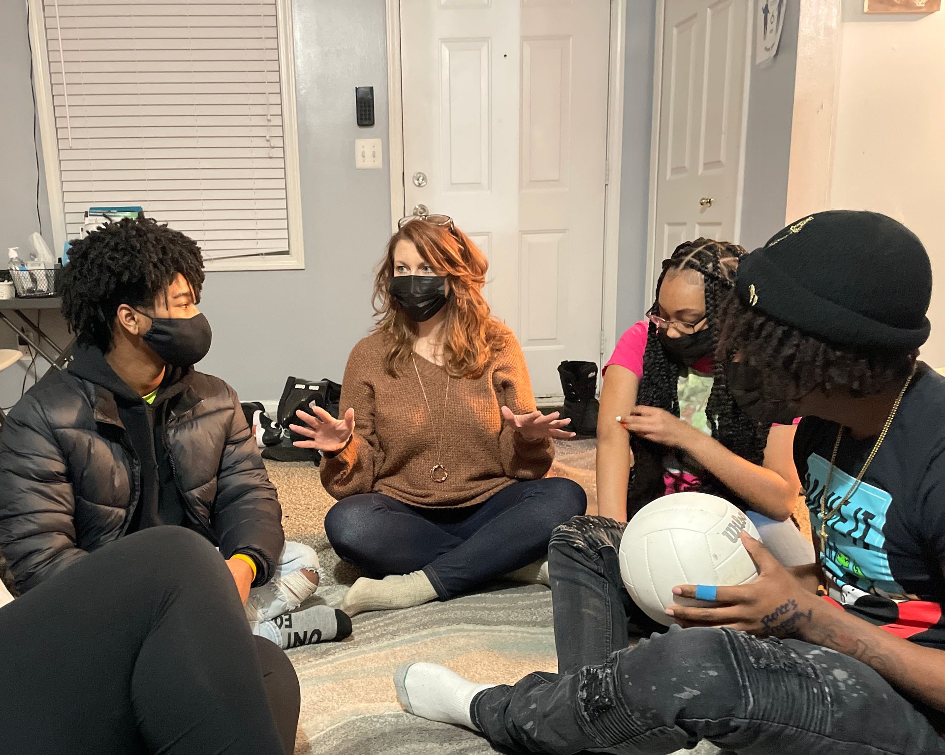 Five masked people sitting in a circle on the floor, talking and holding a white ball indoors