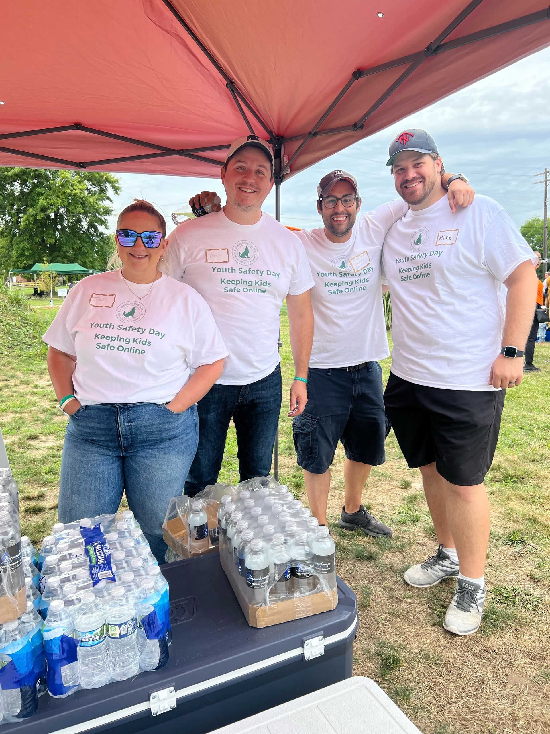 Four volunteers in white shirts stand under a red canopy beside cases of water bottles.