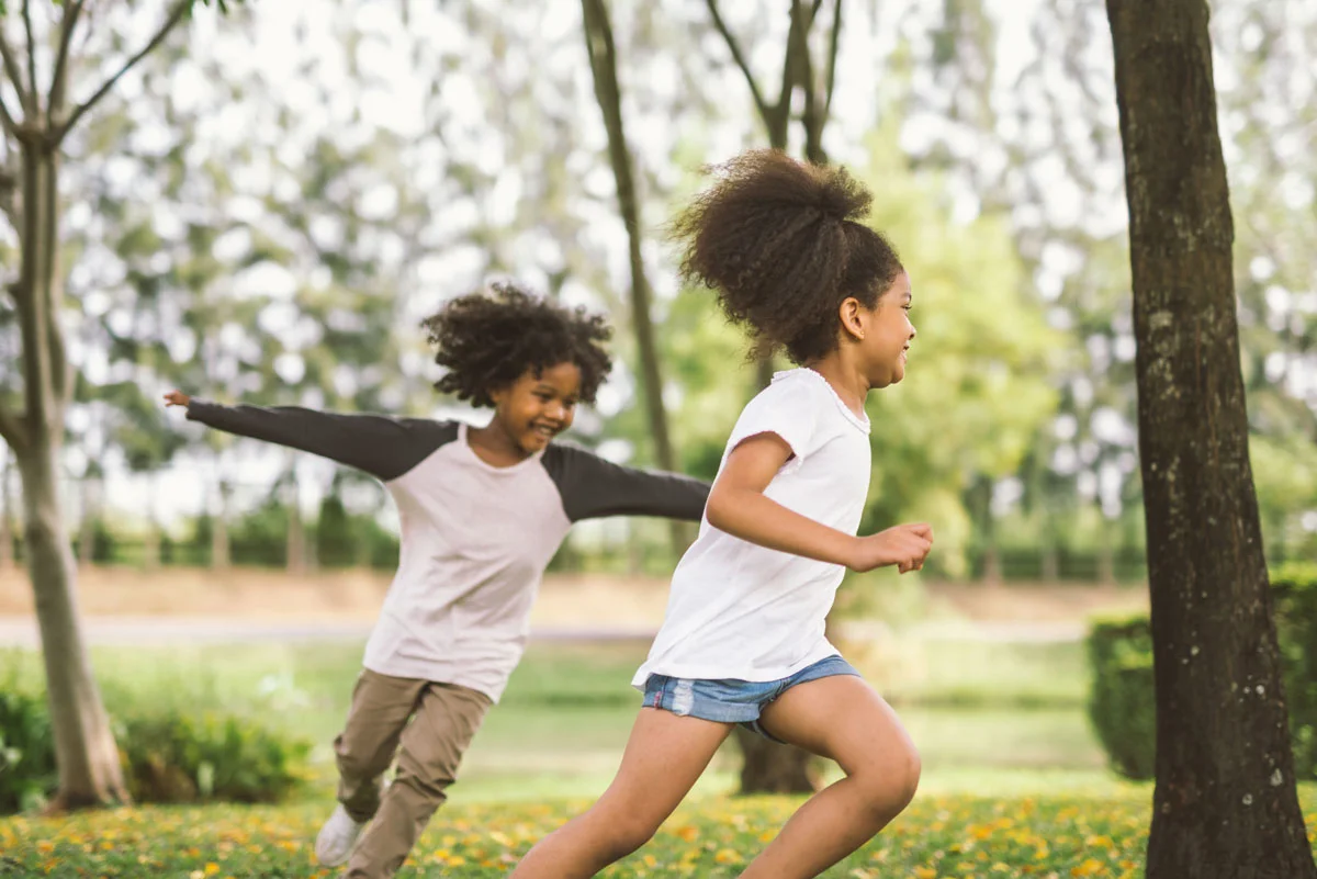 Two children playing tag in a grassy park, smiling and running outdoors.