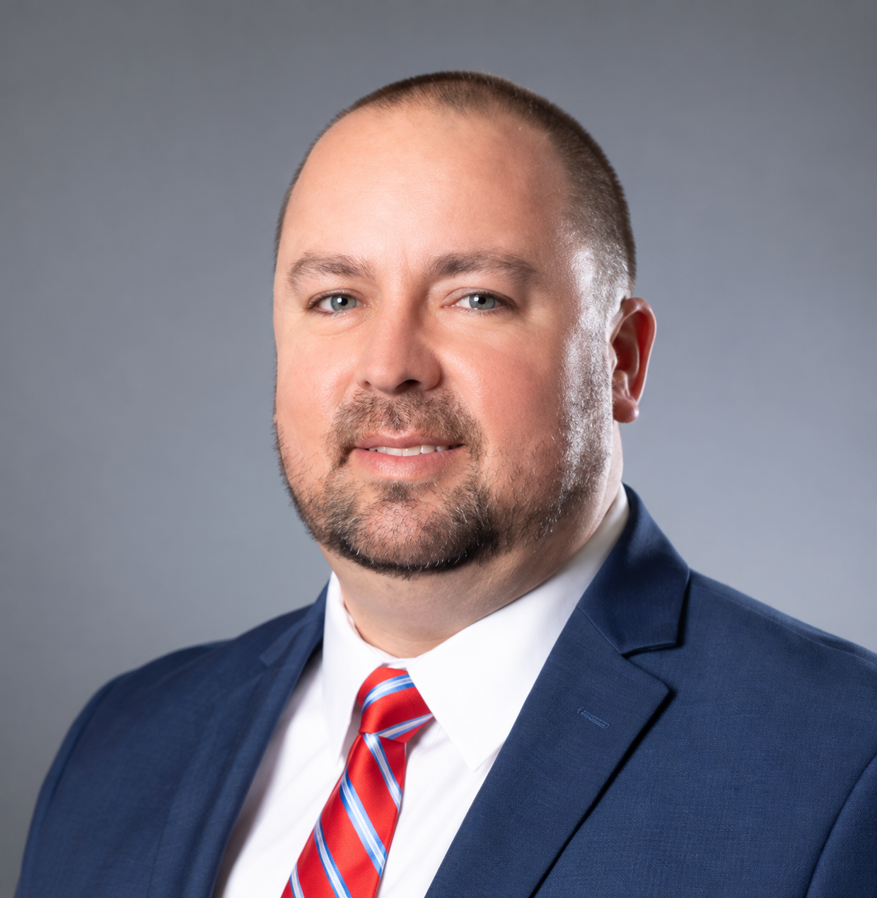 Studio headshot of a man in a blue suit, white shirt, and red striped tie against a gray background