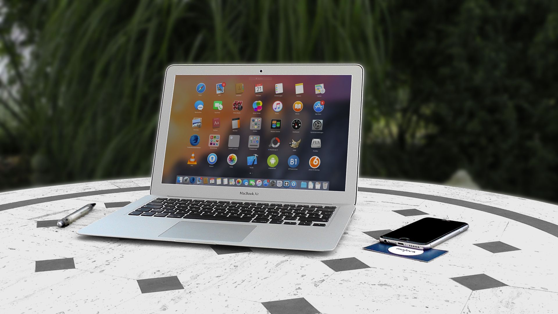 Silver laptop on a white table with a smartphone, stylus, and small accessories, outdoors against greenery