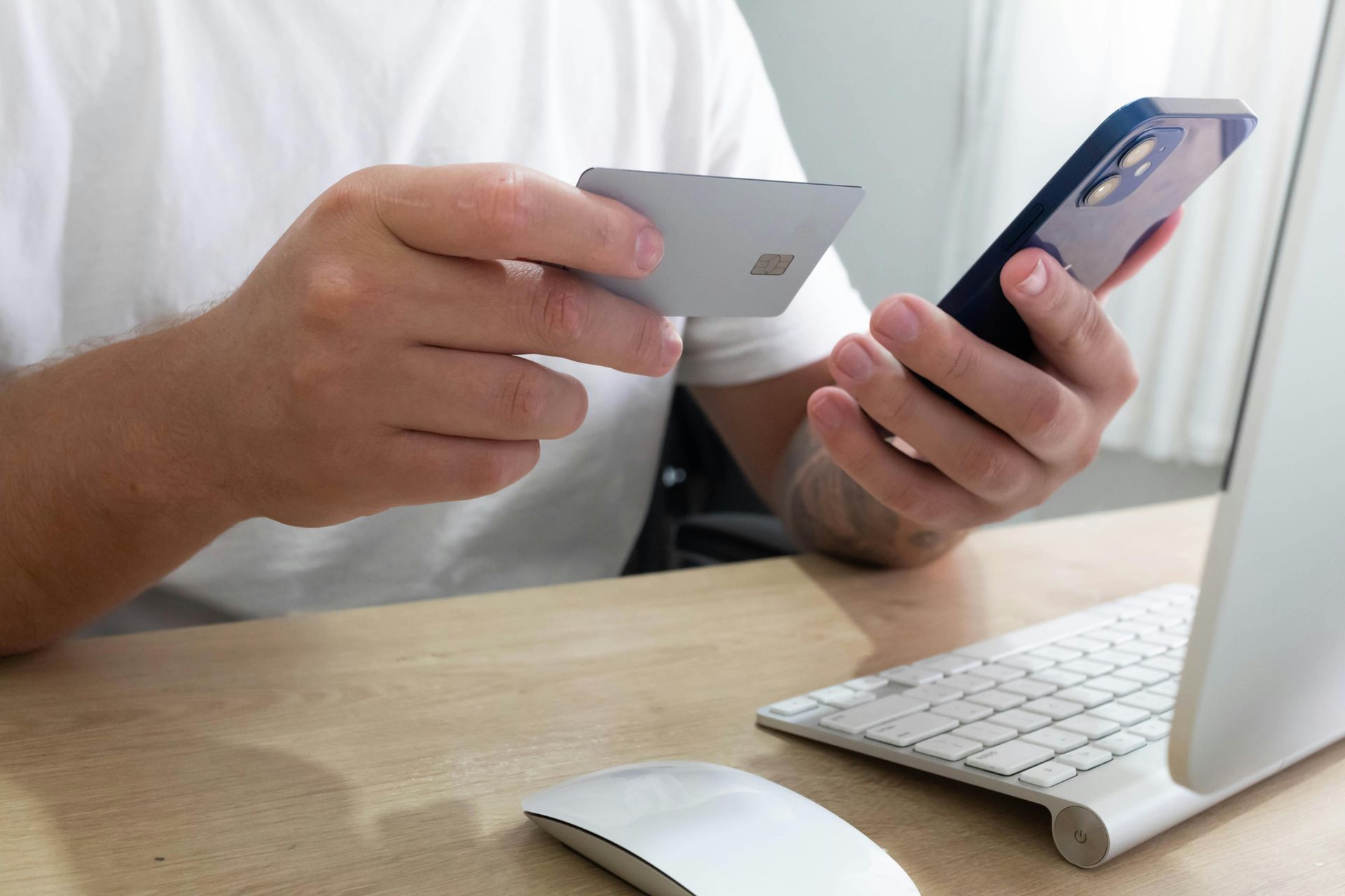 Hands holding a credit card and smartphone at a desk with a keyboard and laptop.