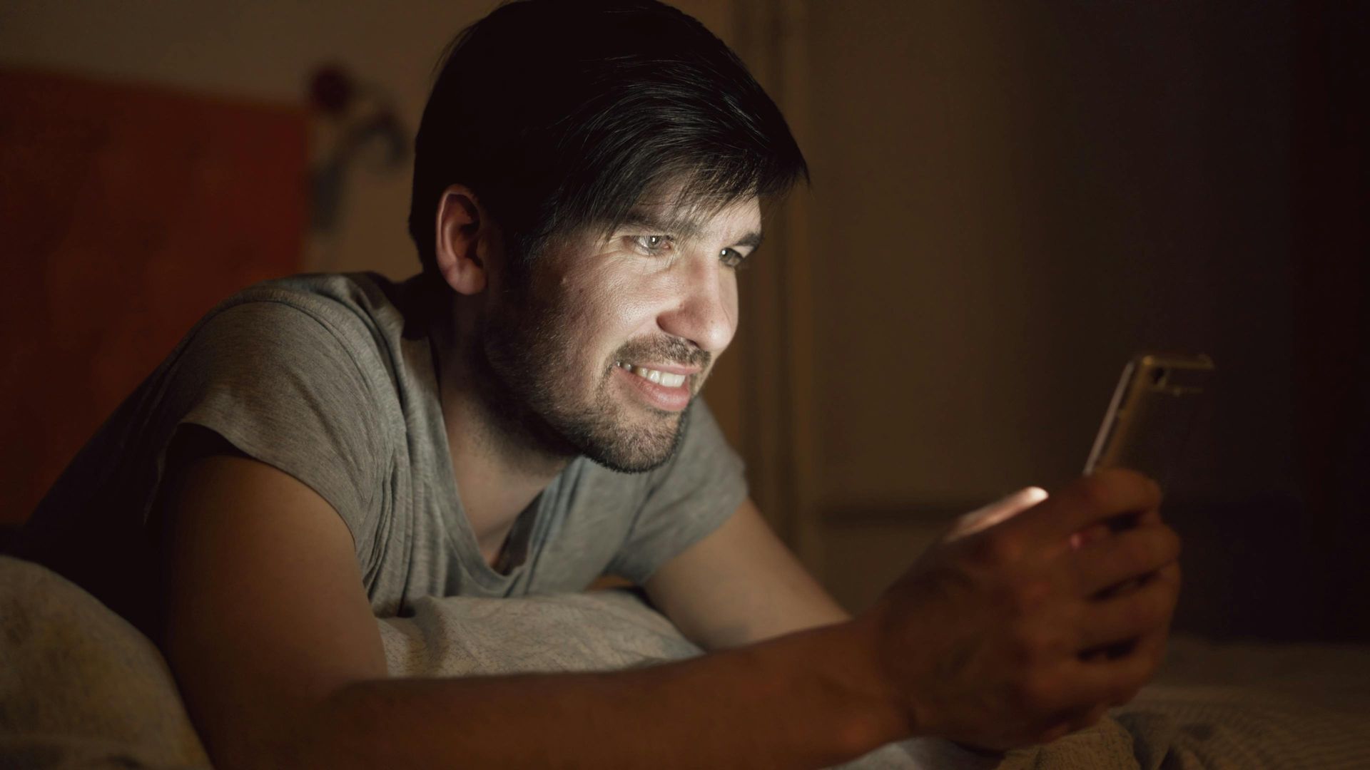 Man lying on a bed, smiling at a glowing phone in the dark.