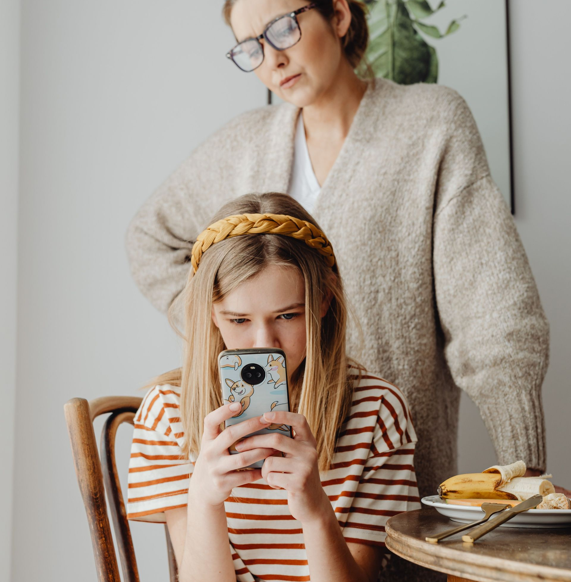Woman and child at a table, child looking at a phone while the woman stands behind her.