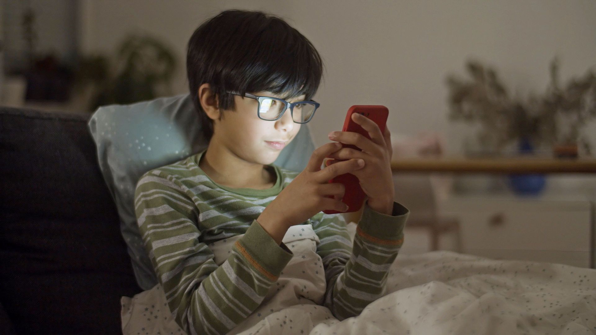 Child in striped pajamas and glasses using a red phone in bed at night