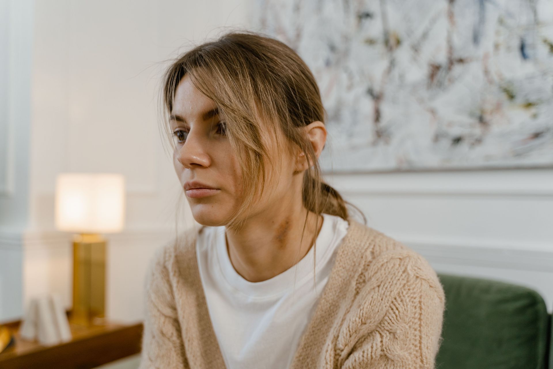 Woman in a beige sweater sitting indoors, looking down beside a lit lamp.