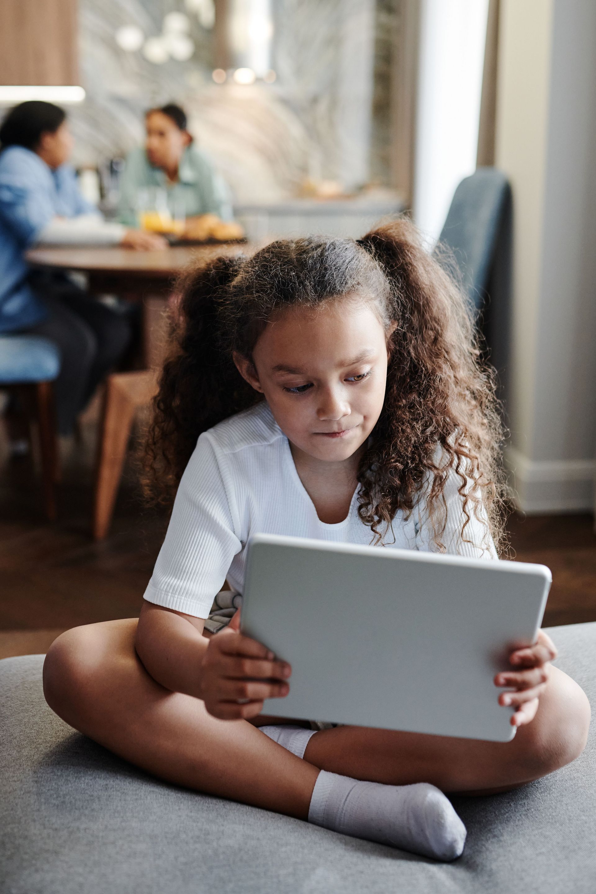 Child sitting on the floor using a tablet in a living room, with two adults blurred in the background