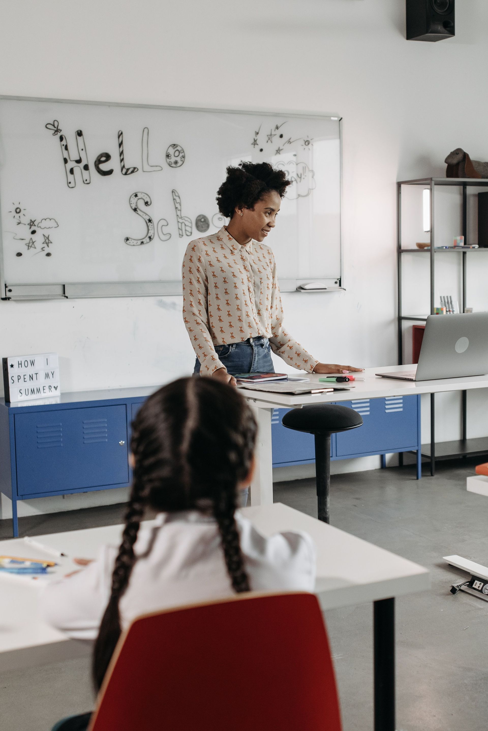 Teacher standing at front of classroom beside laptop and whiteboard, with student seated in foreground
