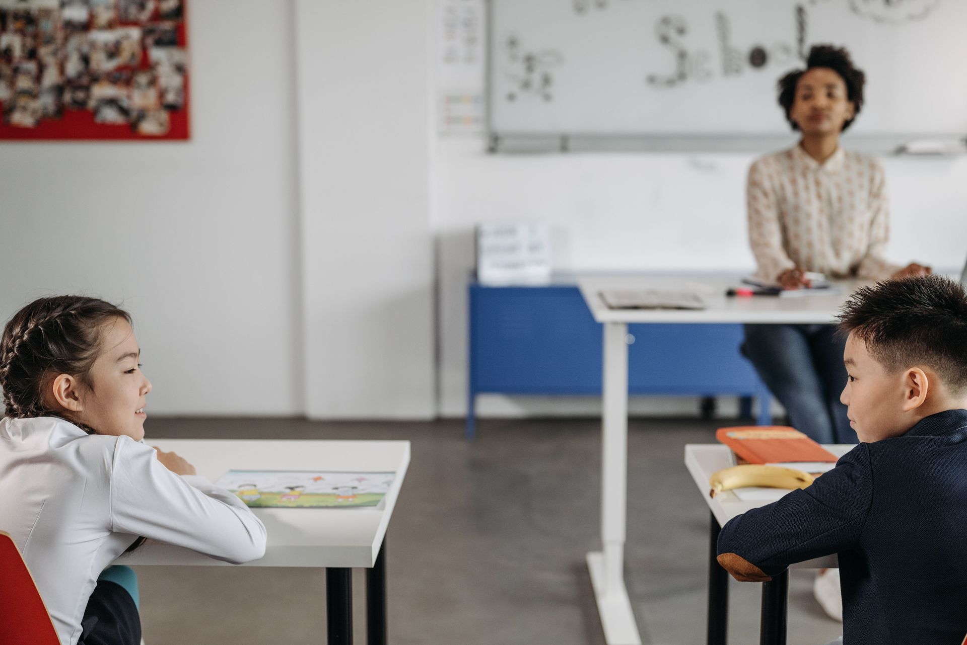 Two students sit at desks in a classroom while a teacher stands at the front.