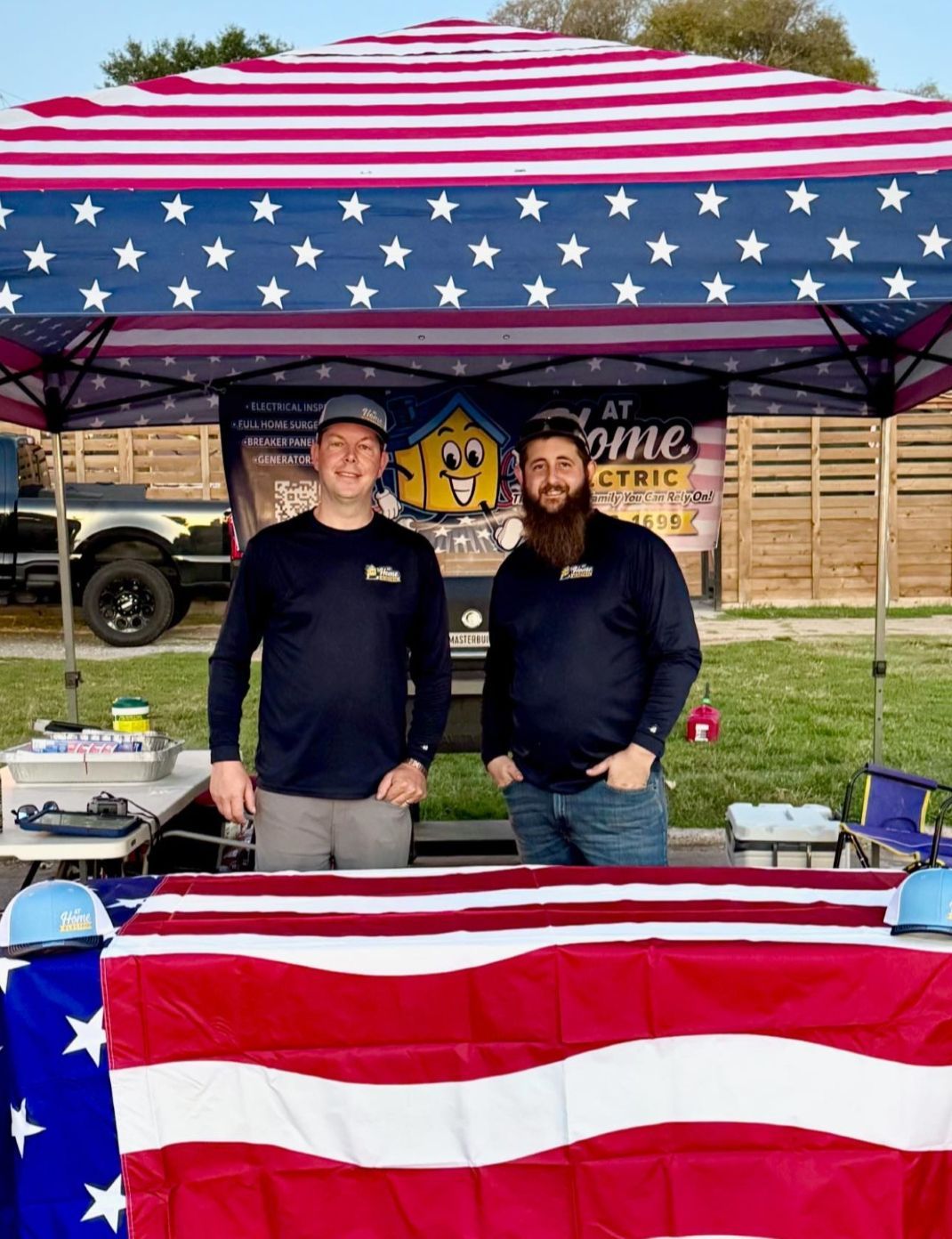 Two people behind a table under a star-spangled tent, with red, white, and blue bunting in front.
