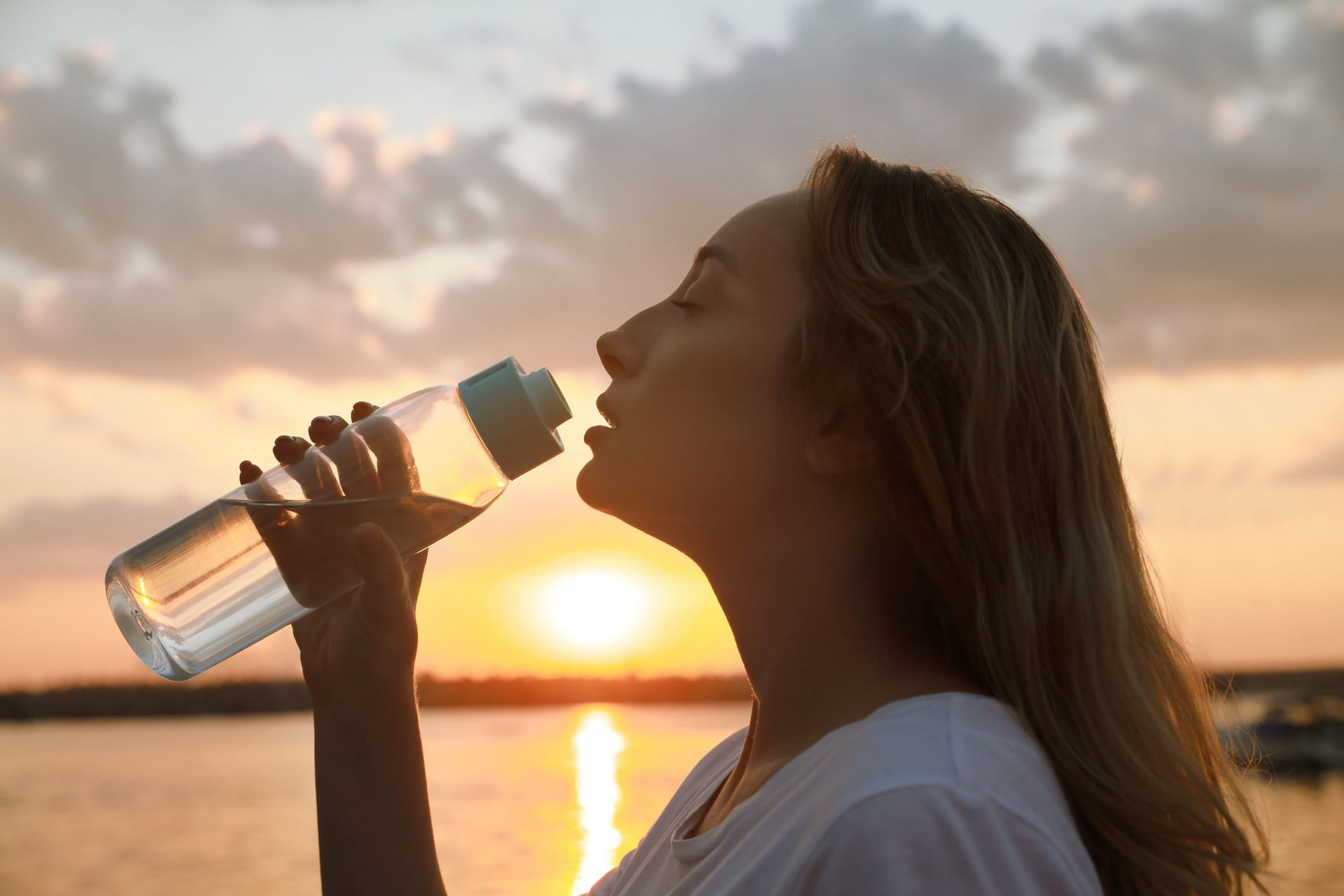 A person drinking from a clear water bottle outdoors against a sunset over water.