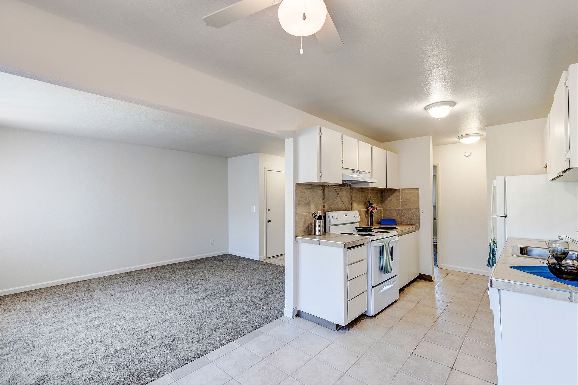 A kitchen with white cabinets , a stove , a refrigerator , and a ceiling fan.