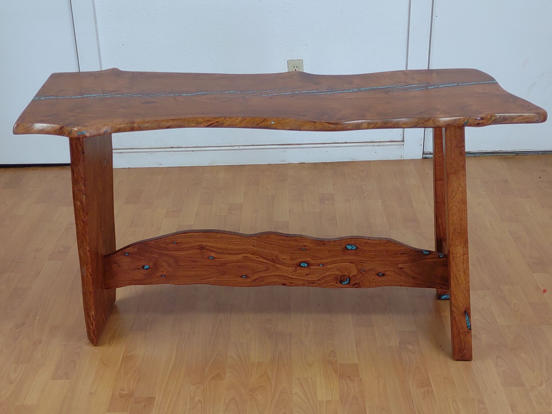 Rustic wooden bench with a natural live-edge top and lower shelf against a plain wall.